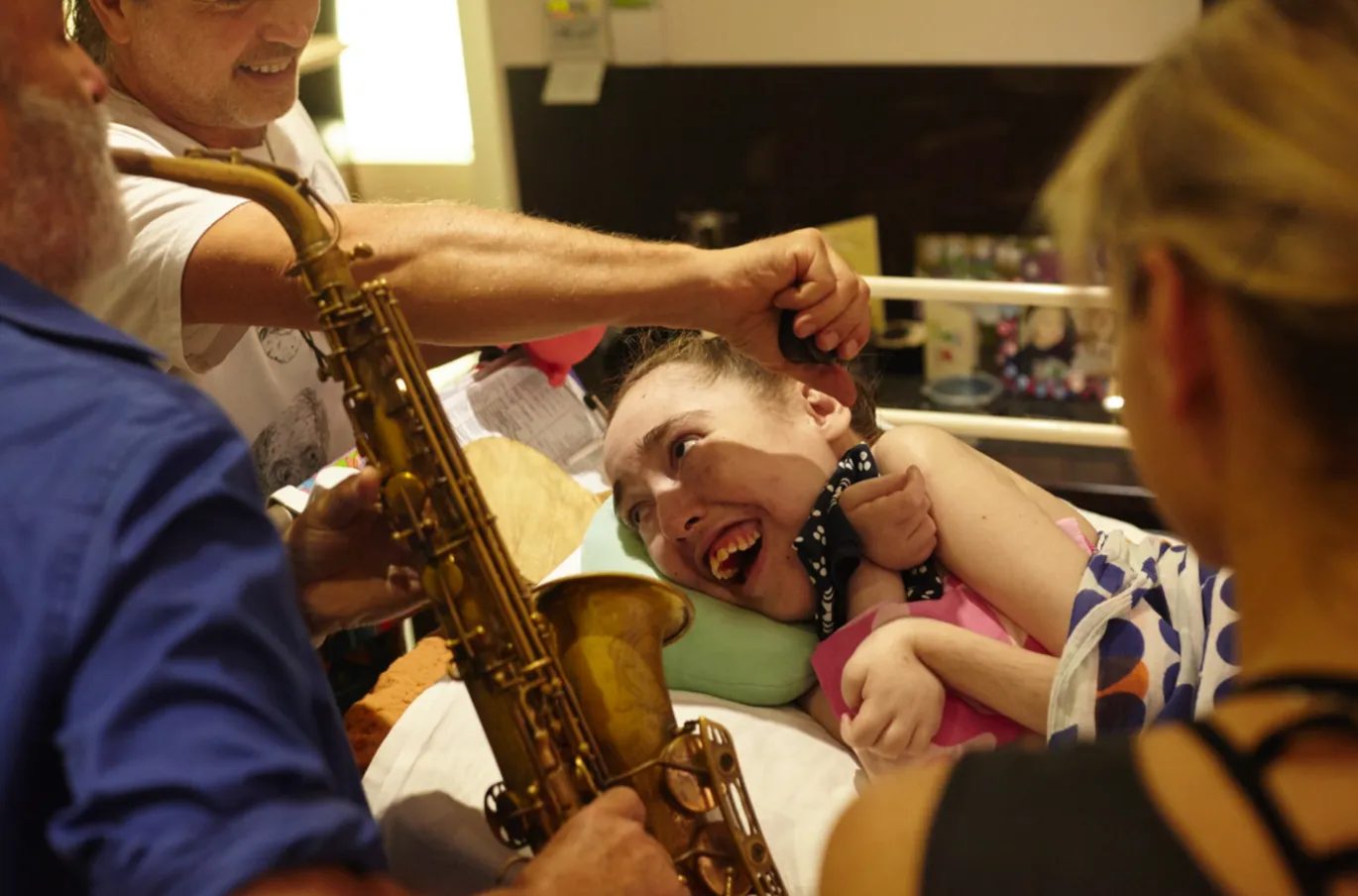 A saxophonist plays at Jess's bedside as she beams with delight, her mother watching on