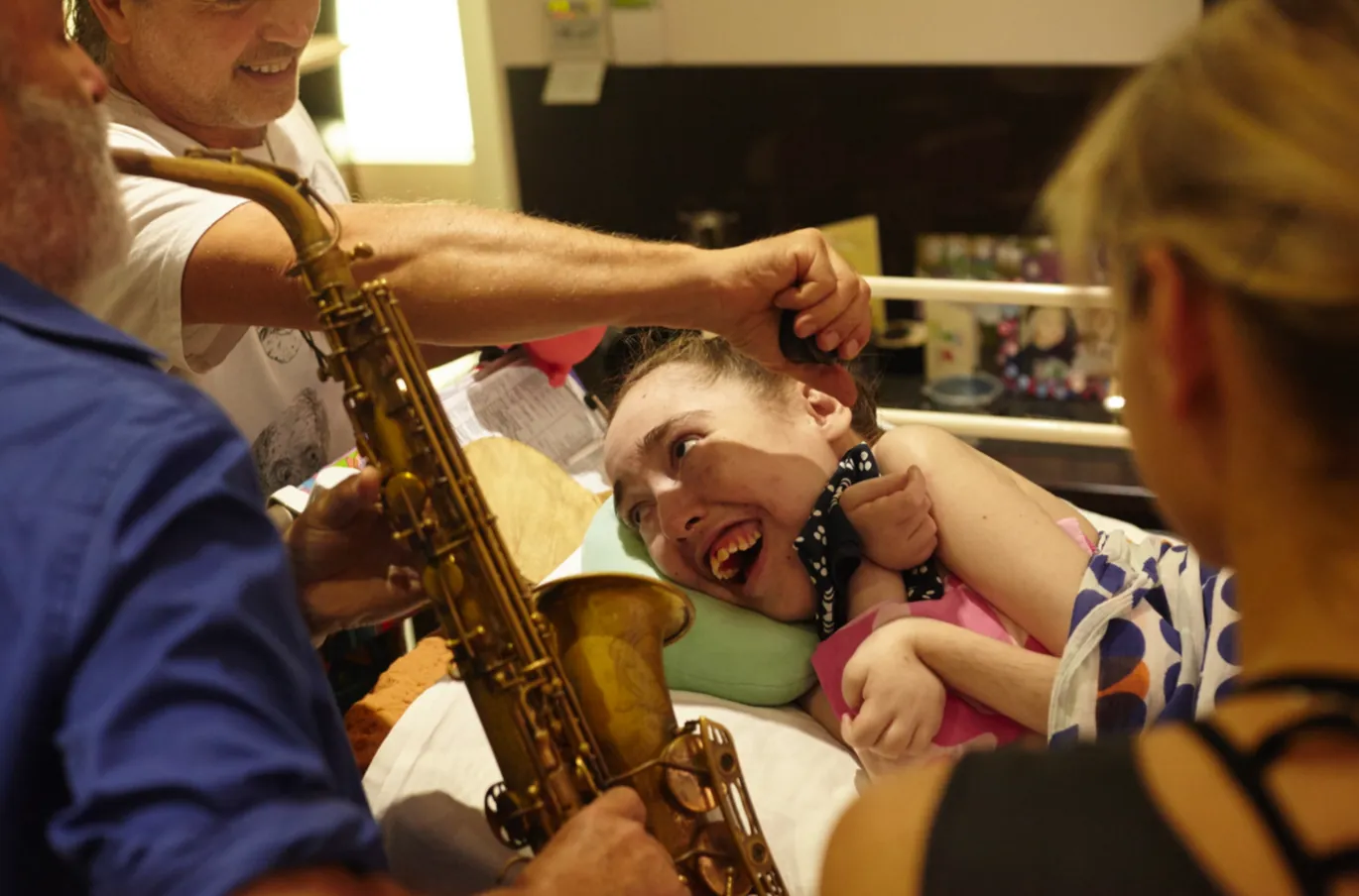 A saxophonist plays at Jess's bedside as she beams with delight, her mother watching on